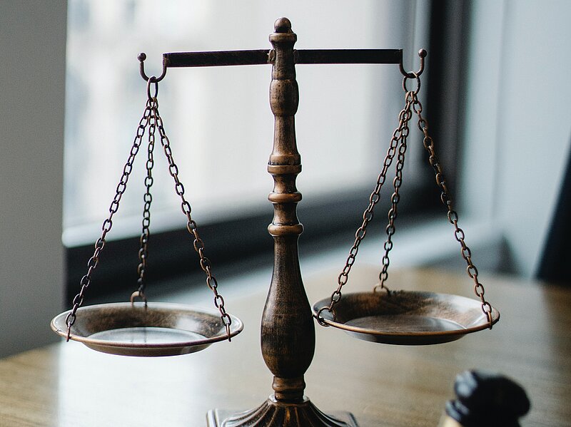 Auf dem Bild ist ein Tisch zuerkennen. Auf diesem steht eine Waage und ein Richterhammer.