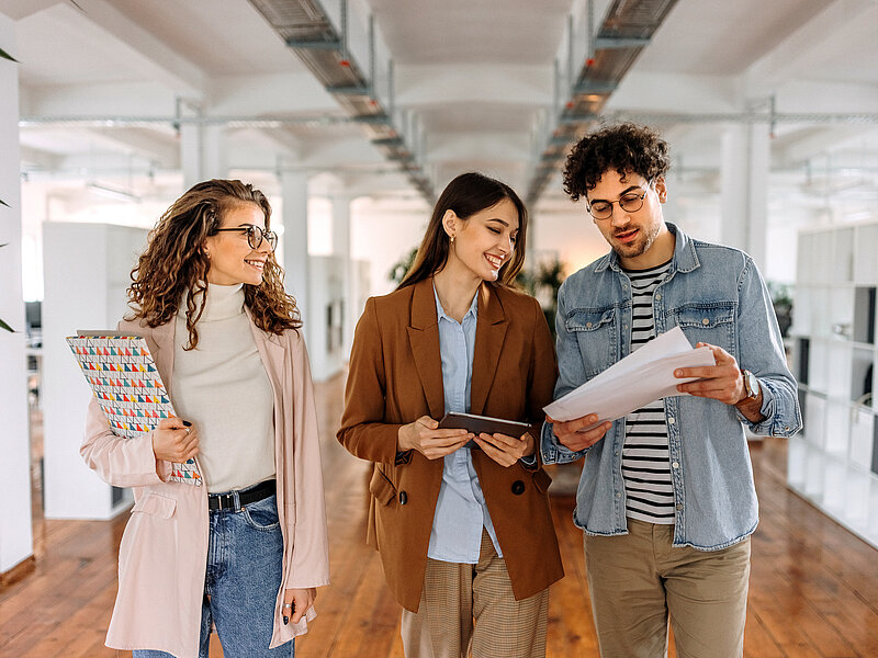Auf dem Bild sieht man drei Personen, welche durchs Büro frontal auf die Kamera zugehen und unterhalten sich dabei. Die Personen halten Dokumente, Papiere und ein ipad in der Hand. 