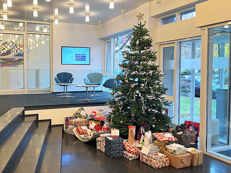 Auf dem Bild ist der Eingangsbereich des kölner Standorts zu sehen. Dort steht der geschmückte Weihnachtsbaum und unter diesen sind ganz viele verpackte Geschenke.